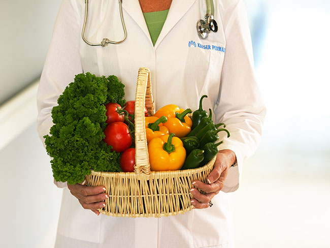 Person wearing Kaiser Permanente whitecoat holding basket containing fresh vegetables